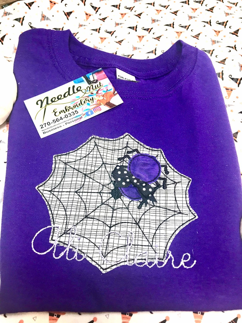 May include: A purple sweatshirt featuring a spider web design with a spider and the name "Claire" embroidered in white. The sweatshirt has a tag from "Needle Nut Embroidery". The background is a Halloween-themed fabric.