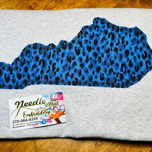 Może przedstawiać: Szara koszulka z niebieskim i czarnym nadrukiem w kształcie stanu Kentucky w stylu lamparta. Koszulka ma wizytówkę Needle Nut Embroidery z numerem telefonu 270-564-0335.