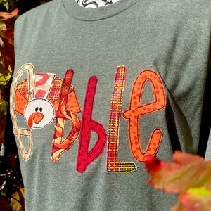 Peut inclure: Un t-shirt vert avec le mot "Gobble" &eacute;crit en lettres de tissu color&eacute;es. Les lettres sont d&eacute;cor&eacute;es de diff&eacute;rents motifs, notamment du plaid, des pois et des tournesols. Le "G" est en forme de dinde avec un bec rouge et un visage blanc.