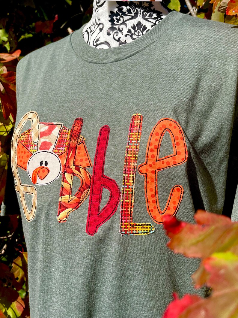 Peut inclure: Un t-shirt vert avec le mot "Gobble" &eacute;crit en lettres de tissu color&eacute;es. Les lettres sont d&eacute;cor&eacute;es de diff&eacute;rents motifs, notamment du plaid, des pois et des tournesols. Le "G" est en forme de dinde avec un bec rouge et un visage blanc.