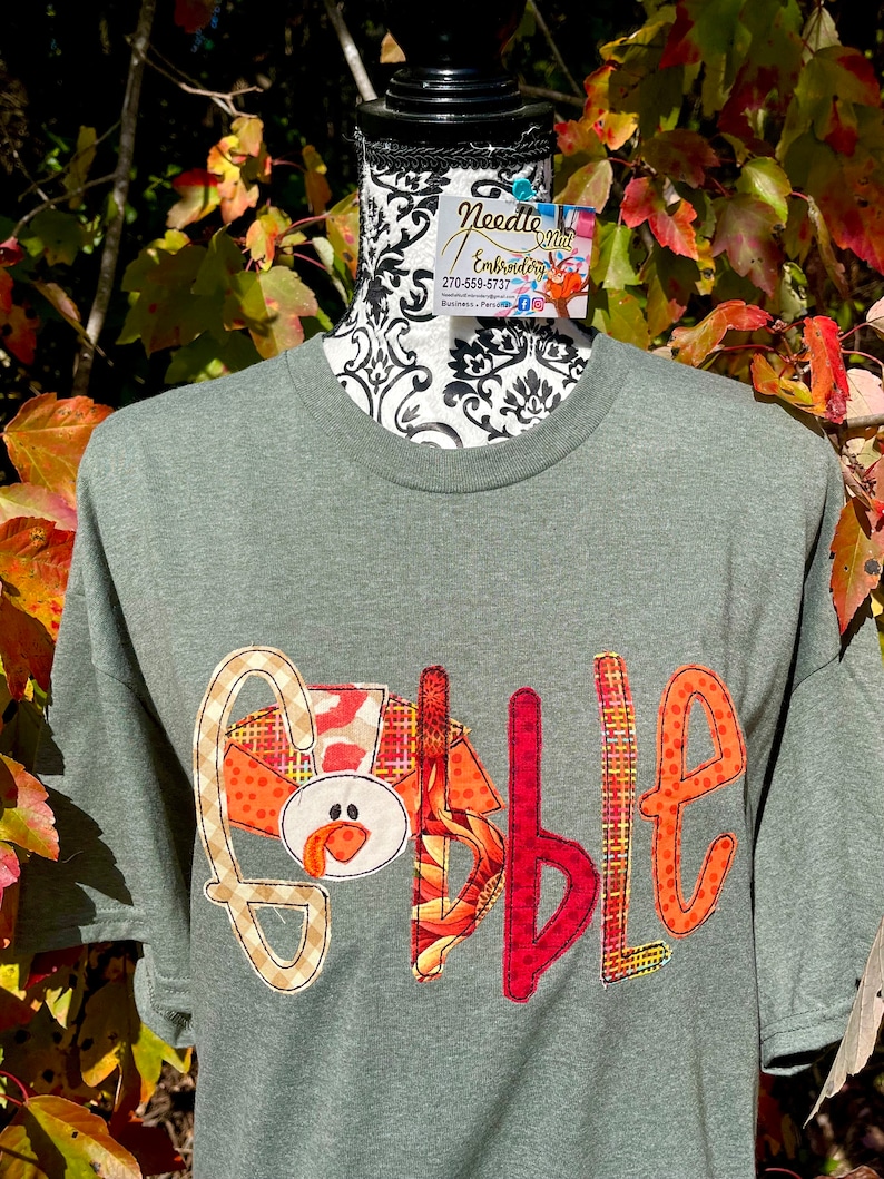 Peut inclure: Un t-shirt vert avec un motif en patchwork qui &eacute;pelle "Gobble" avec une t&ecirc;te de dinde dans le "G".