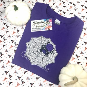 May include: A purple crew neck sweatshirt featuring a spider web design with a spider. The spider is black with purple polka dots. The text "Cinderella" is embroidered on the shirt. Two white pumpkins are on either side of the shirt.