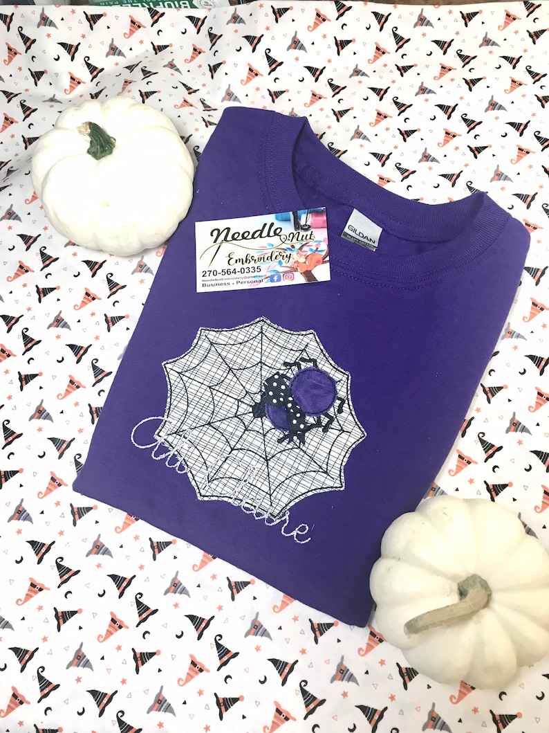 May include: A purple crew neck sweatshirt featuring a spider web design with a spider. The spider is black with purple polka dots. The text "Cinderella" is embroidered on the shirt. Two white pumpkins are on either side of the shirt.