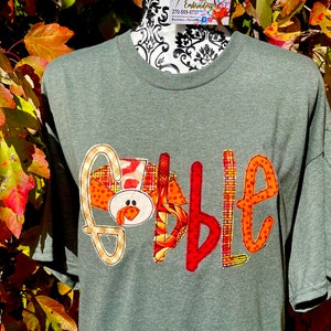Peut inclure: Un t-shirt vert avec le mot "Gobble" &eacute;crit en patchs de tissu color&eacute;s. Le "G" est en forme de dinde avec un corps &agrave; motifs rouge, orange et jaune et une t&ecirc;te brune avec une barbichette rouge.
