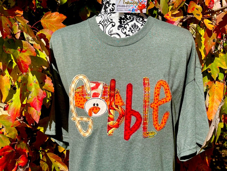 Peut inclure: Un t-shirt vert avec le mot "Gobble" &eacute;crit en patchs de tissu color&eacute;s. Le "G" est en forme de dinde avec un corps &agrave; motifs rouge, orange et jaune et une t&ecirc;te brune avec une barbichette rouge.