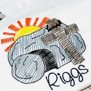 May include: White t-shirt with an embroidered design of a stone tomb, a cross, and a rising sun. The name "Riggs" is embroidered below the design.