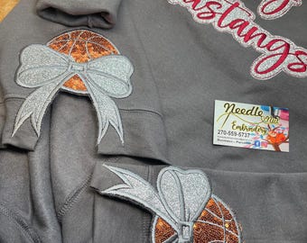 May include: Gray sweatshirt with the words "Lady Mustangs" embroidered in pink and silver glitter. The sweatshirt has a basketball with a silver glitter bow on each sleeve. The text "Emma Kate" is embroidered on the left sleeve. A business card for "Needle Nut Embroidery" is visible in the bottom right corner.