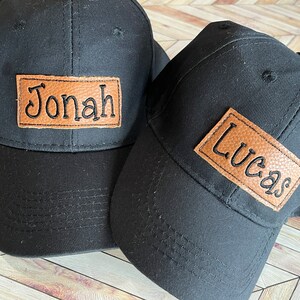 Op de afbeelding: Twee zwarte baseballpetten met bruine leren patches. De patches hebben de namen "Jonah" en "Lucas" erop geborduurd in zwarte draad.