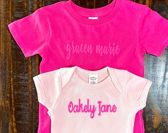 May include: Two pink and white short-sleeved shirts with names printed on them. The pink shirt says "gracen marie" and the white shirt says "Oakely Jane".