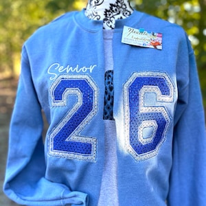 Könnte beinhalten: Hellblaues Sweatshirt mit dem Wort "Senior" in weißer Schrift. Große, paillettenbesetzte "26" Ziffern in Blau und Silber sind auf der Vorderseite. Das Sweatshirt wird auf einer Schaufensterpuppe präsentiert.