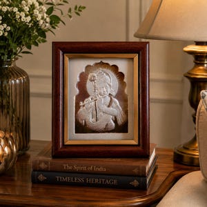 May include: A silver relief of a deity in a dark wood frame, on a stack of books. The relief shows a figure with a halo, playing a flute. The books are titled "The Spirit of India" and "Timeless Heritage".