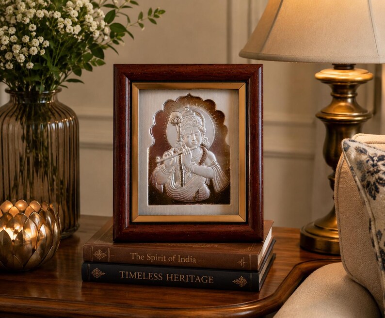 May include: A silver relief of a deity in a dark wood frame, on a stack of books. The relief shows a figure with a halo, playing a flute. The books are titled "The Spirit of India" and "Timeless Heritage".
