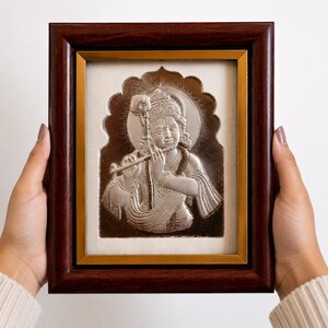May include: A framed silver relief of a figure playing a flute, set against a cream background. The frame is dark brown with a gold inner border. The artwork features intricate details and a halo.