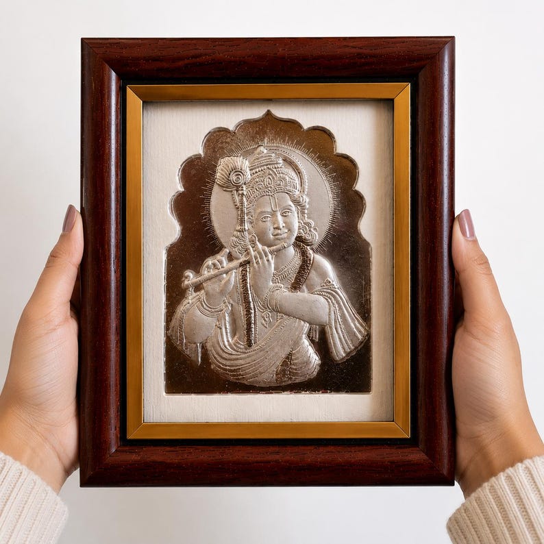 May include: A framed silver relief of a figure playing a flute, set against a cream background. The frame is dark brown with a gold inner border. The artwork features intricate details and a halo.
