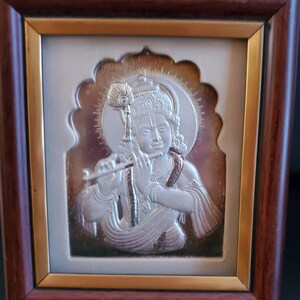 May include: A framed silver relief sculpture of a deity playing a flute. The sculpture is set against a bronze background and framed in a dark wood frame with a gold inner border. The deity is depicted with a halo.