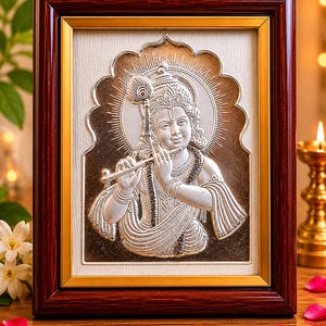 May include: A framed silver relief sculpture of a deity playing a flute. The artwork is set against a cream background and framed in dark brown wood with a gold inner border. A peacock feather adorns the deity's head.