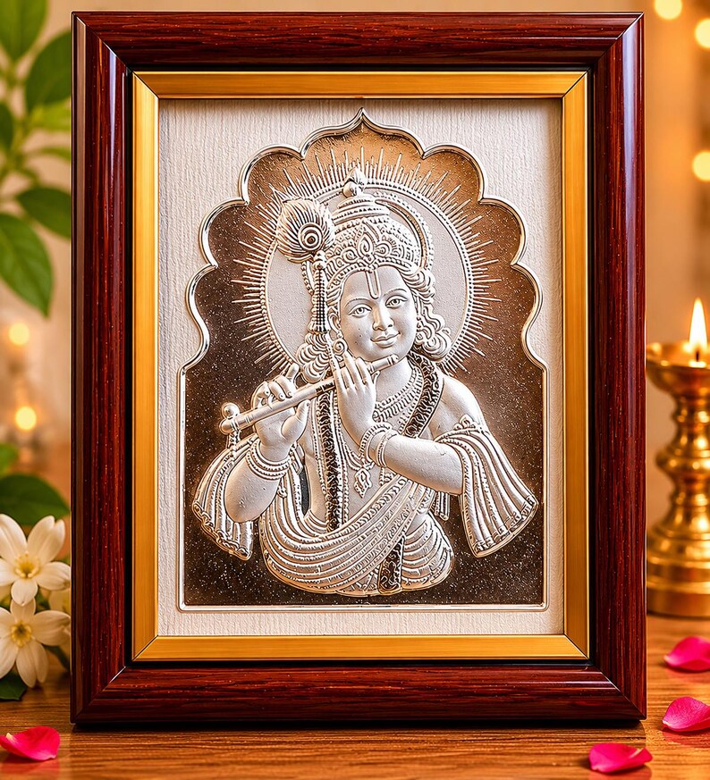 May include: A framed silver relief sculpture of a deity playing a flute. The artwork is set against a cream background and framed in dark brown wood with a gold inner border. A peacock feather adorns the deity's head.