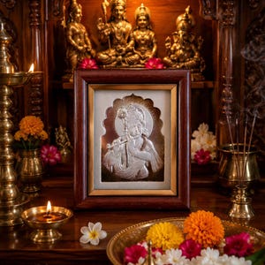 May include: A framed silver relief of a deity, displayed on a wooden altar with other religious statues and candles. The relief depicts a figure playing a flute. A brass bowl of flowers and a lit candle are in the foreground.