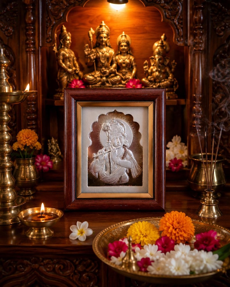 May include: A framed silver relief of a deity, displayed on a wooden altar with other religious statues and candles. The relief depicts a figure playing a flute. A brass bowl of flowers and a lit candle are in the foreground.