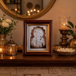 May include: A framed silver relief of a deity playing a flute, set against a gold background. The frame is dark brown. The image is part of a home decor display, with candles, flowers, and a decorative mirror.
