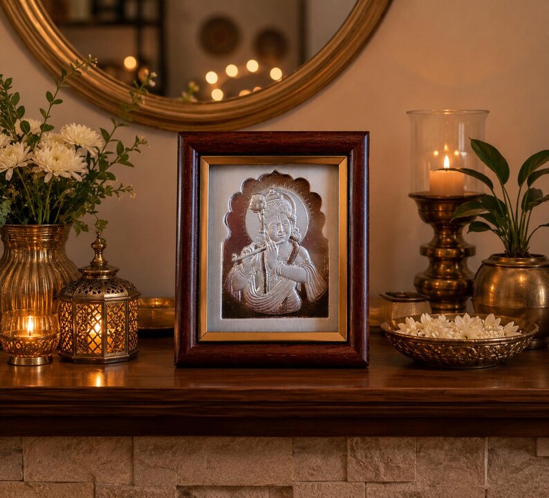May include: A framed silver relief of a deity playing a flute, set against a gold background. The frame is dark brown. The image is part of a home decor display, with candles, flowers, and a decorative mirror.
