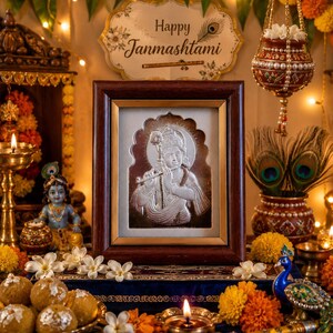 May include: A framed silver relief of a deity playing a flute, surrounded by festive decorations. The scene includes marigold flowers, candles, and a sign reading "Happy Janmashtami".