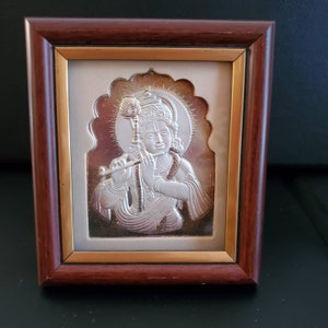 May include: A framed silver relief sculpture of a deity playing a flute. The artwork is set against a light background and framed in a dark brown wood frame with a gold inner border. The deity wears jewellery and has a halo.