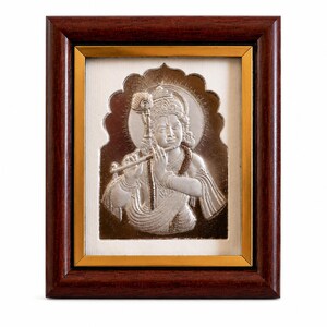 May include: A framed silver relief sculpture of a figure playing a flute, set against a white background. The artwork is framed in a dark brown wood frame with a gold inner border. The figure has a halo.