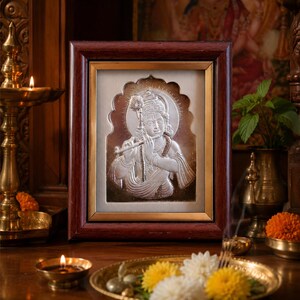May include: A framed silver relief depicting a deity playing a flute. The frame is dark brown with a gold inner border. The image is set against a cream background. Candles, flowers, and brass religious items surround the frame.