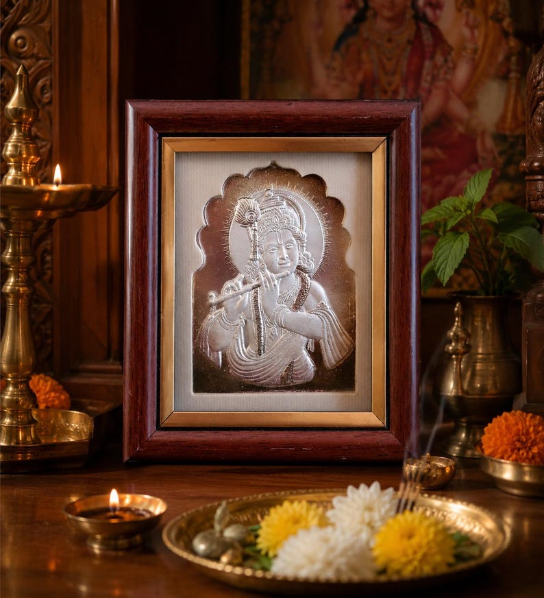 May include: A framed silver relief depicting a deity playing a flute. The frame is dark brown with a gold inner border. The image is set against a cream background. Candles, flowers, and brass religious items surround the frame.