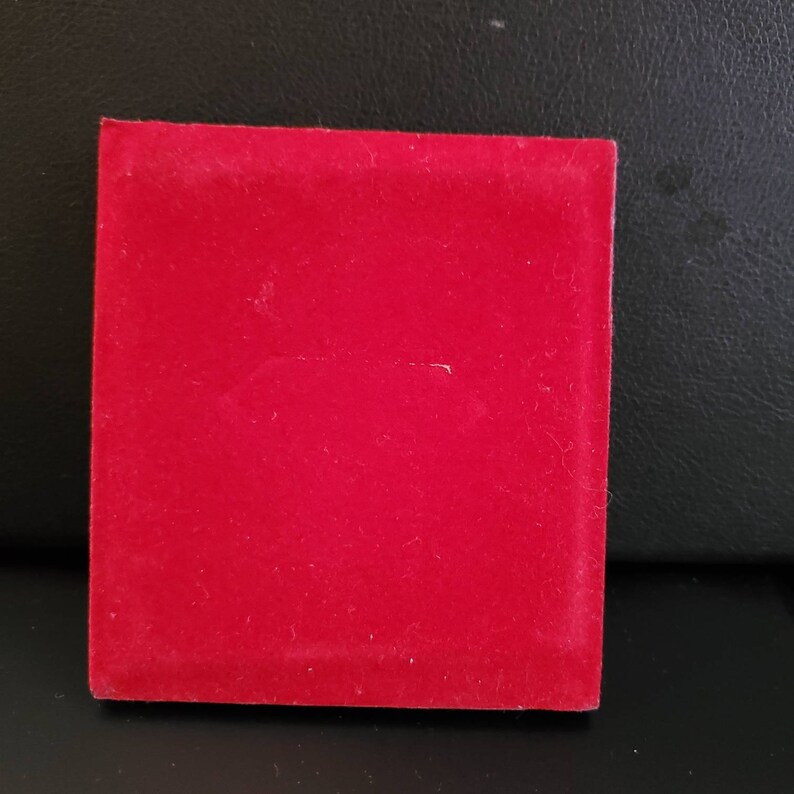 May include: A square, red velvet box. The box is a solid, vibrant red colour. The velvet material has a soft, slightly worn appearance. The box is sitting on a black surface.