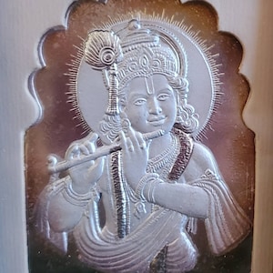 May include: A silver relief sculpture of a deity playing a flute, set within an ornate, arched border. The figure has a halo and a peacock feather. The background has a gradient effect, transitioning from dark to light.