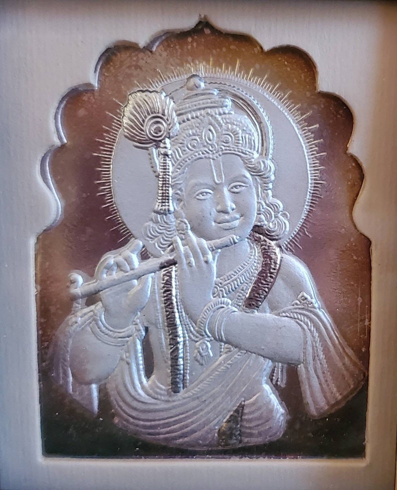 May include: A silver relief sculpture of a deity playing a flute, set within an ornate, arched border. The figure has a halo and a peacock feather. The background has a gradient effect, transitioning from dark to light.