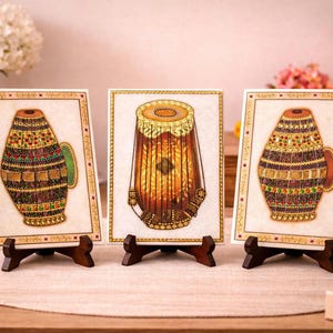 Op de afbeelding: Drie ingelijste kunstwerken met gedetailleerde illustraties van Indiase muziekinstrumenten. De kunstwerken tonen een tabla-drum en twee decoratieve vaten, allemaal versierd met ingewikkelde patronen en levendige kleuren, op houten standaards.