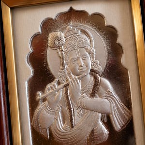 May include: A framed silver relief sculpture of a deity playing a flute. The sculpture has detailed features, including a crown, jewellery, and flowing garments. The frame is a combination of gold and dark brown wood.