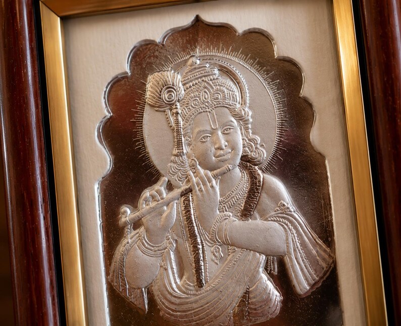 May include: A framed silver relief sculpture of a deity playing a flute. The sculpture has detailed features, including a crown, jewellery, and flowing garments. The frame is a combination of gold and dark brown wood.