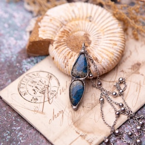 May include: A pendant necklace featuring two teardrop-shaped blue and gray stones set in a silver-toned metal frame. The pendant is attached to a delicate silver-toned chain with small spherical beads spaced along the length of the chain.