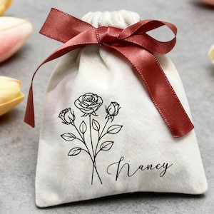 Könnte beinhalten: Ein weißer Samtbeutel mit einer schwarzen Rosenillustration und dem Namen "Nancy" in Schreibschrift. Der Beutel ist mit einem dunkelroten Satinband gebunden. Tulpen und ein silberner Ring mit einem großen Stein sind im Hintergrund.