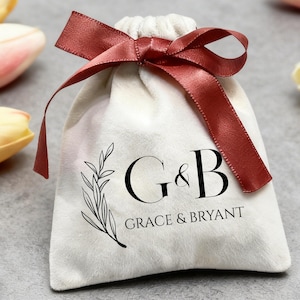 Op de afbeelding: Een kleine, crèmekleurige fluwelen geschenktas met een donkerrode satijnen strik. De tas heeft de initialen "G&B" en de woorden "GRACE & BRYANT" in zwart, samen met een zwarte botanische illustratie. Een diamanten ring en tulpen staan op de achtergrond.