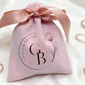 Puede incluir: Una bolsa de joyería de terciopelo rosa claro con un lazo a juego, con un logotipo circular con el texto "GRACE & BRYAN" y un diseño floral. Se ven varias joyas, incluyendo anillos y un collar.