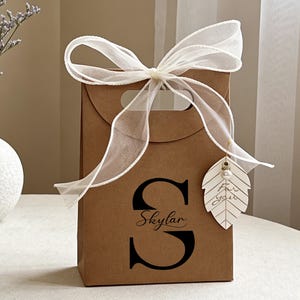 May include: A brown gift bag tied with a white ribbon bow and a leaf-shaped tag. The bag features a handle and the letter "S" with the name "Skylar". The leaf tag has the words "for you" and a pearl.