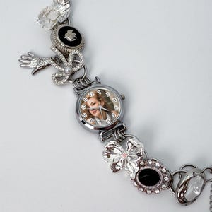 May include: Silver-toned watch bracelet with a round watch face featuring a woman's portrait. The bracelet is adorned with various decorative elements, including bows, a hand, and floral accents. The watch face has a diameter of approximately 1 inch.