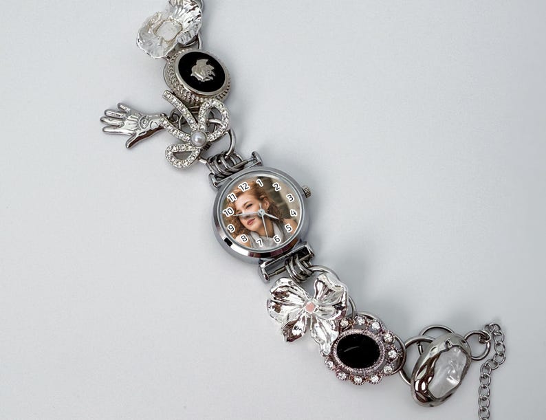 May include: Silver-toned watch bracelet with a round watch face featuring a woman's portrait. The bracelet is adorned with various decorative elements, including bows, a hand, and floral accents. The watch face has a diameter of approximately 1 inch.