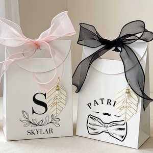 May include: Two white gift bags with decorative bows. One has a pink bow, the other a black bow. Each bag has a leaf-shaped charm and personalized text. One says "Skylar" and the other says "Patri".