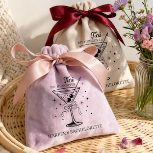 Peut inclure: Deux petits sacs à cordon, un lavande et un crème, sont posés sur un plateau en osier. Le sac lavande a un ruban rose et un dessin de verre à martini avec le texte "Tini Bit Hungover" et "Harper's Bachelorette". Le sac crème a un ruban bordeaux et un texte similaire.