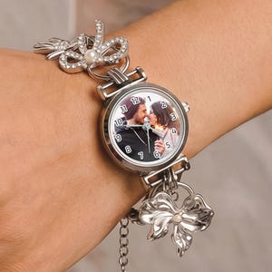 May include: Silver-toned wristwatch with a round face featuring a couple kissing. The watch has a silver band with bow and pearl accents. The watch face displays the time and a photo of a couple. The watch is on a wrist.