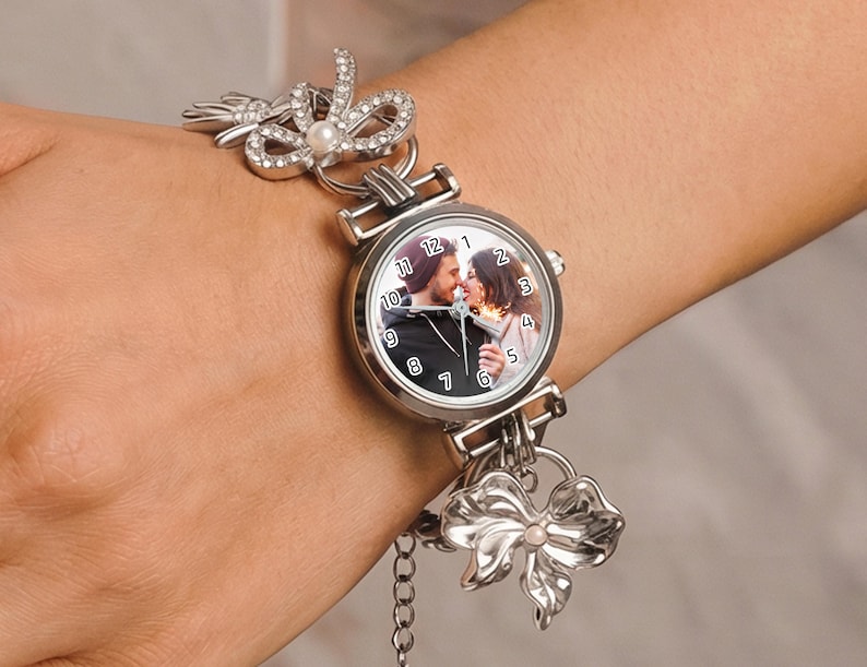 May include: Silver-toned wristwatch with a round face featuring a couple kissing. The watch has a silver band with bow and pearl accents. The watch face displays the time and a photo of a couple. The watch is on a wrist.
