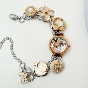 May include: A personalized watch bracelet with a gold-tone watch face displaying a couple's photo. The bracelet features decorative charms in gold and white, including a flower, a butterfly, and a starburst design. The watch band is silver-toned.