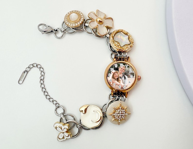 May include: A personalized watch bracelet with a gold-tone watch face displaying a couple's photo. The bracelet features decorative charms in gold and white, including a flower, a butterfly, and a starburst design. The watch band is silver-toned.