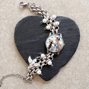 May include: Silver-toned watch bracelet with a chain link design, adorned with faux pearls and a small angel charm. The watch face displays a photo of a couple. The bracelet is resting on a black heart-shaped stone.
